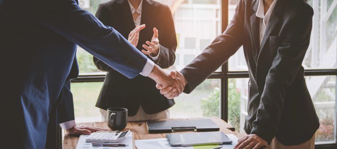 Two business people shaking hands across table with third person applauding