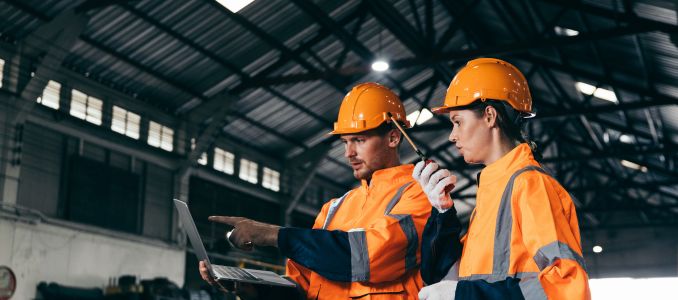 Two warehouse or industrial workers wearing bright orange safety jackets and hard hats collaborate inside a large facility.
