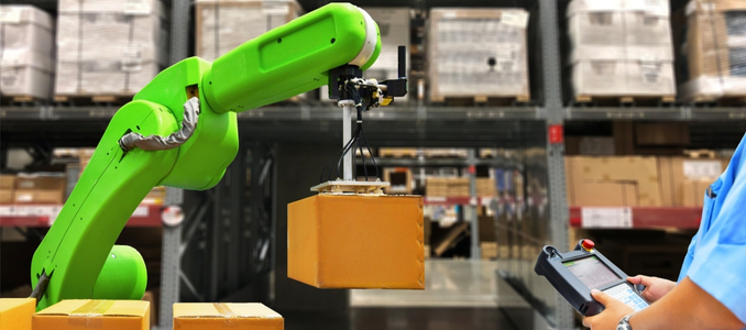 A bright green robotic arm lifts a cardboard box using a suction attachment inside a warehouse, while a worker in a blue shirt controls it using a handheld terminal in the foreground.