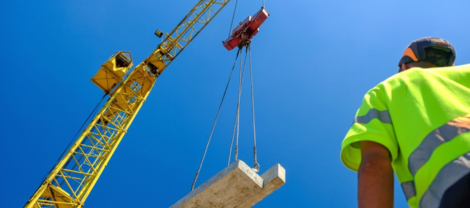 A construction worker looking up at a yellow crane lifting a heavy concrete slab against a clear blue sky.