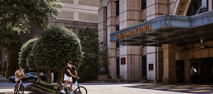 Photo courtesy of Four Seasons Hotel showing the grand entrance with prominent golden signage above the marquee as guests ride bicycles along the driveway in front.