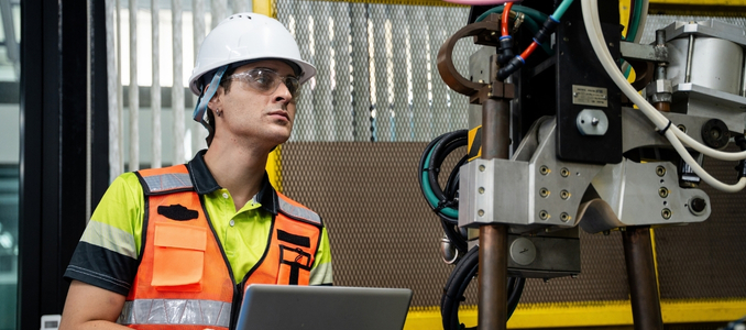 An industrial worker wearing a white hard hat, safety glasses, and an orange safety vest inspecting heavy machinery while holding a laptop.