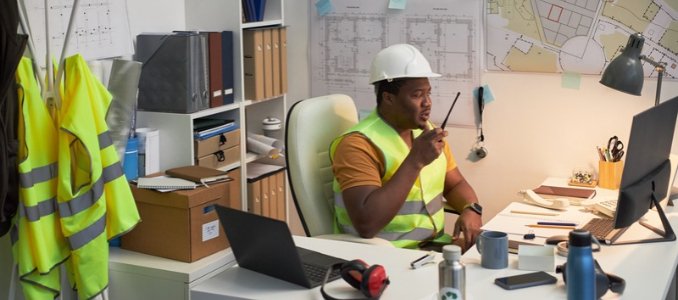 A site manager in a hard hat and safety vest speaking into a walkie-talkie at a site office desk filled with computers and architectural blueprints.