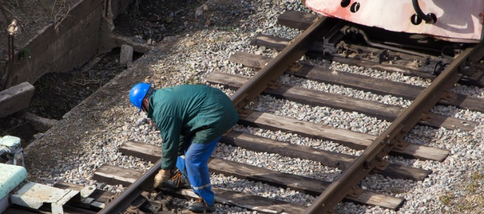 An NTSB investigator wearing a blue hard hat and green jacket inspecting the metal tracks near a train car on a railway line.