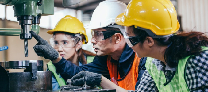 Three factory workers in hard hats and safety goggles receiving hands-on equipment safety training around a vertical drill press machine.