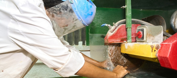 A worker wearing a protective face shield uses a wet-cutting saw with a continuous water spray to cut stone or tile, suppressing silica dust.