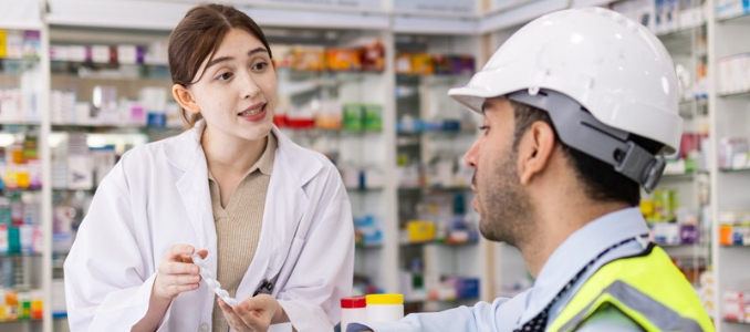 A pharmacist in a lab coat speaks with a worker wearing a hard hat and safety vest.