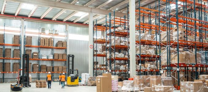 Two workers walk through a warehouse lined with tall shelving, stacked pallets and boxes.