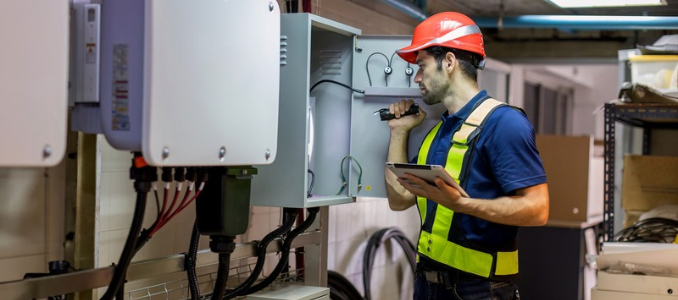 An electrician wearing a red hard hat and safety vest uses a flashlight and tablet to inspect an open electrical panel box.
