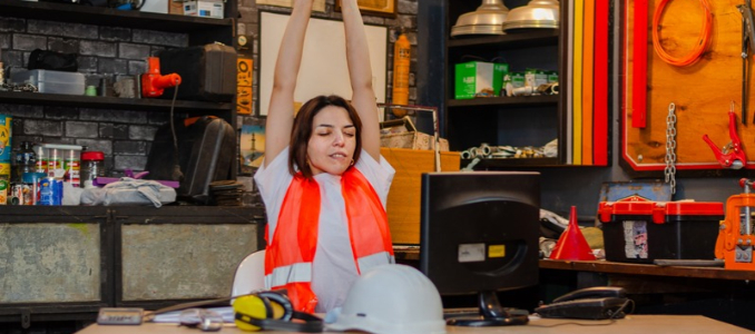 A woman in a safety vest stretches her arms overhead while sitting at a desk in a workshop with tools and a computer monitor.