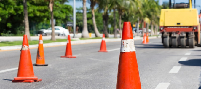 A row of orange traffic cones blocks off a lane on an asphalt road during construction, with a large yellow steamroller parked in the background and palm trees lining the street.