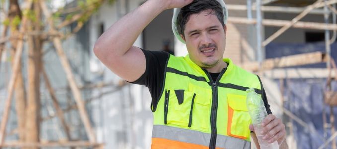 Construction worker in a high-visibility vest wipes his forehead while holding a water bottle at a job site.