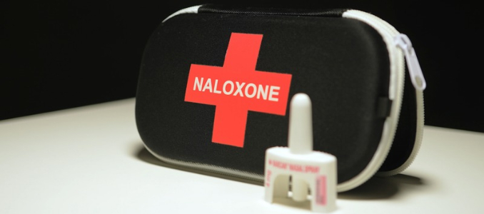 A Naloxone nasal spray device standing in front of a black and red emergency response kit.