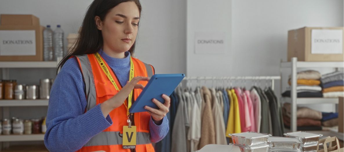 Volunteer in an orange reflective vest uses a tablet beside donated food, clothing and boxes.