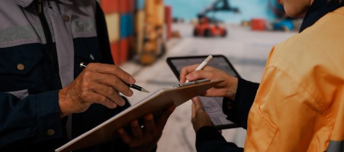 Two workers review a clipboard and tablet in a shipping container yard.
