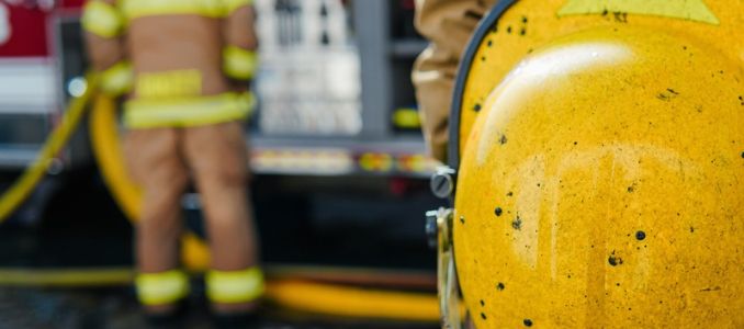 Close-up of a yellow firefighter helmet with a firefighter, fire engine and hoses in the background.