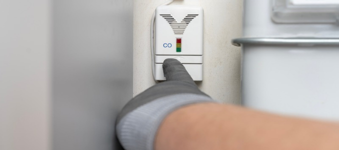 A gloved hand pressing the test button on a wall-mounted carbon monoxide detector.
