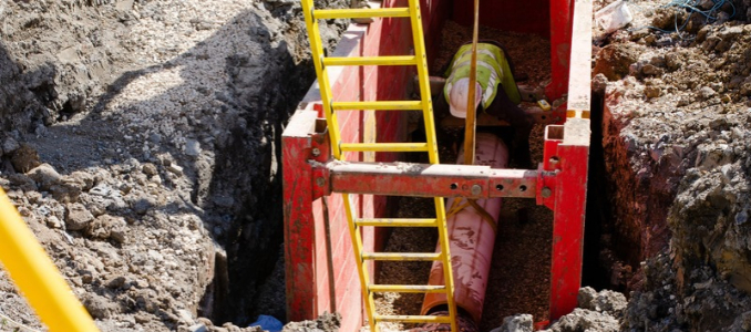 A worker wearing a safety vest and hard hat working inside a trench protected by a red trench box, with a yellow ladder extending down into the excavation site.