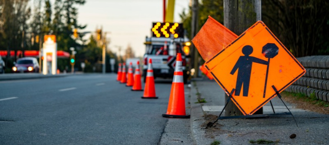 An orange road construction sign displaying a worker figure along a roadside lined with traffic cones and a construction truck with illuminated flashing arrows.