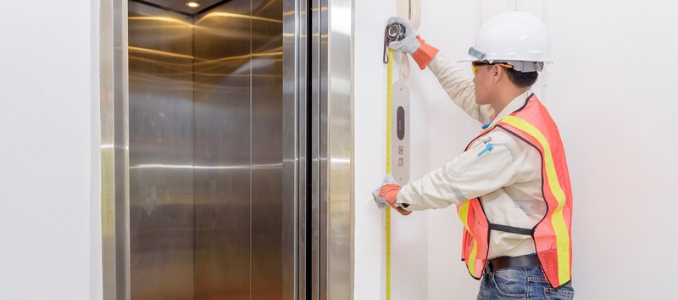 A technician wearing a white hard hat and high-visibility safety vest using a tape measure next to open elevator doors.