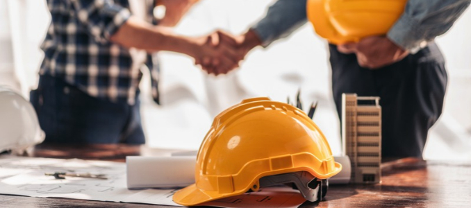Two professionals shaking hands over a table with yellow hard hats, blueprints, and a architectural model in the foreground.