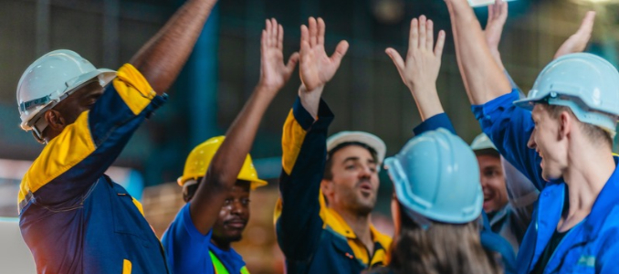 A team of industrial workers wearing hard hats celebrating with high-fives in a factory setting.