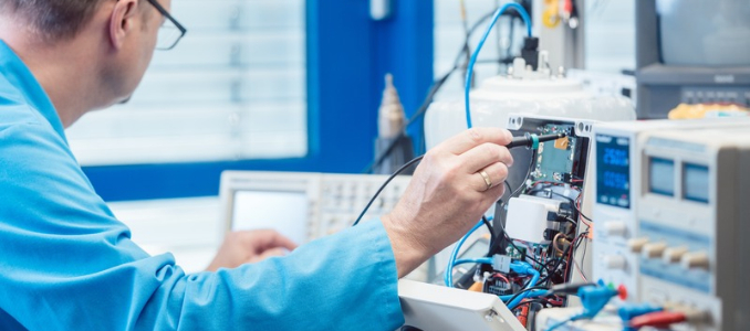 An technician in a blue lab coat using a probe to test circuit boards and electrical components in a testing laboratory.