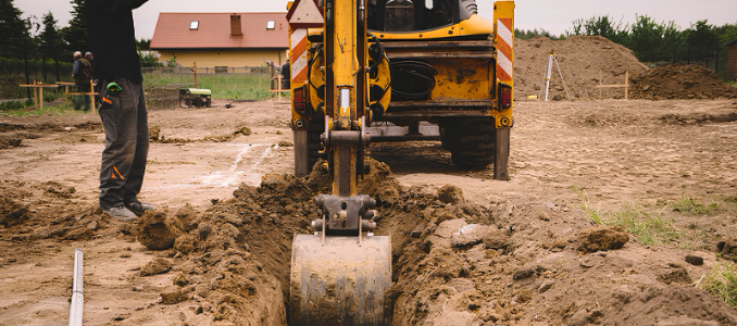 Backhoe bucket digging a narrow trench at a construction site, with a worker standing nearby.
