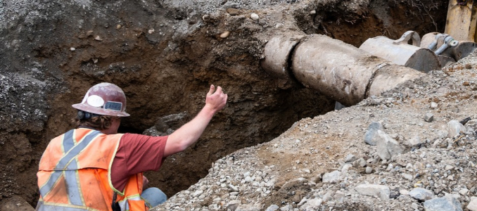 Construction worker wearing a hard hat and safety vest works in a deep excavation beside a large underground pipe.