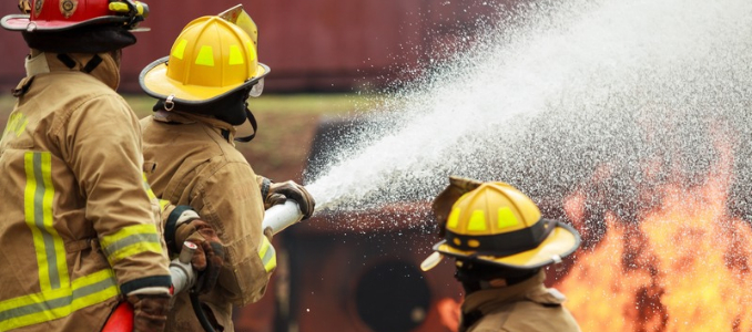 Firefighters in protective gear spray foam onto flames during a firefighting operation.