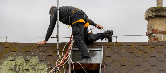 Worker wearing a safety harness climbs onto a rooftop with ropes and equipment.