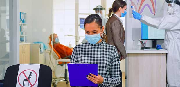 person wearing a facemask in black and white checkered shirt writing on purple clipboard in dentist office. various kinds of ppe protocol can be seen, including a sign indicating no one can sit in a chair next to the person in the checkered shirt and a receptionist in full PPE in the background taking a patient