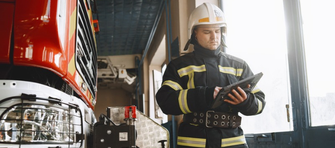 A firefighter wearing full protective turn-out gear and a helmet operating a digital tablet beside a fire truck inside a fire station bay.