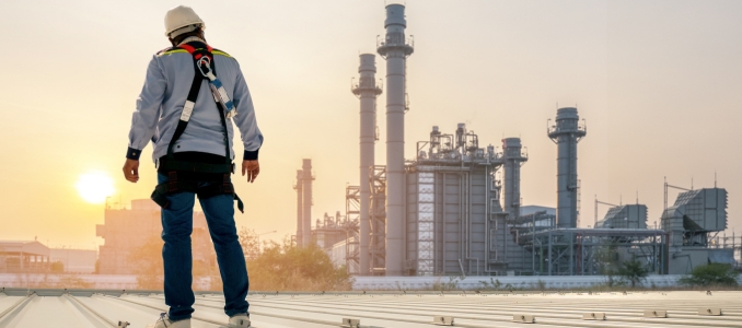 A lone worker, outfitted with safety gear, stands on a rooftop, surveying a vast industrial complex as the sun rises