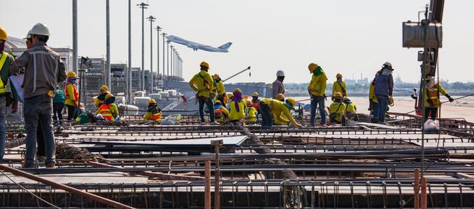 A large team of construction workers wearing hard hats and high-visibility vests work on a rebar-covered structure, with an airplane taking off in the background near an airport terminal.