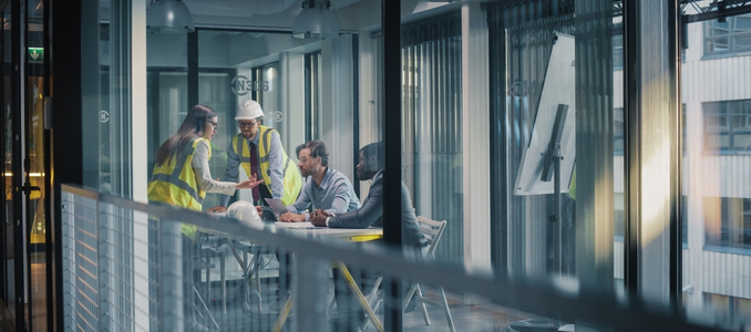 A group of four professionals - including construction engineers in safety vests and office executives—gathered around a table in a modern glass conference room for a meeting.