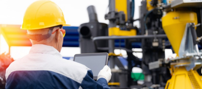 Industrial worker reviews digital operating information on a tablet beside manufacturing equipment.