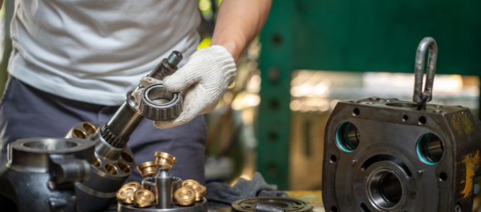 A gloved technician holds a bearing beside the disassembled parts of a hydraulic pump on a workshop bench.