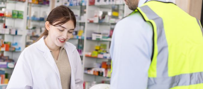Female pharmacist in lab coat providing medical consultation to industrial worker in safety vest at pharmacy