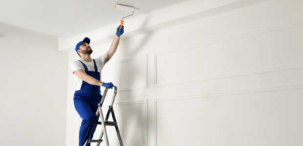 a person in blue overalls painting a ceiling white while standing on a ladder