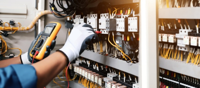 An electrician using a digital multimeter to test wiring and components inside an electrical control panel.