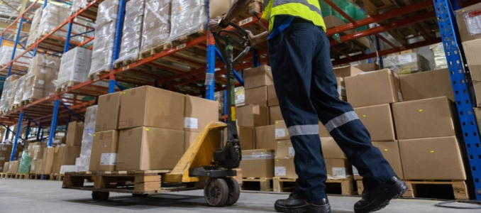Warehouse worker in a high-visibility vest operating a pallet jack among stacked boxes.