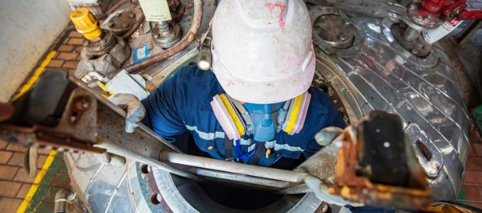 Worker wearing a hard hat, respirator and gloves climbs a ladder through an opening in an industrial tank.