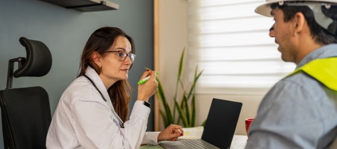 Clinician wearing a white coat and stethoscope speaks with a worker wearing a hard hat and high-visibility vest.