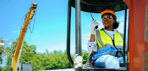 woman in backhoe holding a walkie talkie while looking to the left against a blue sky