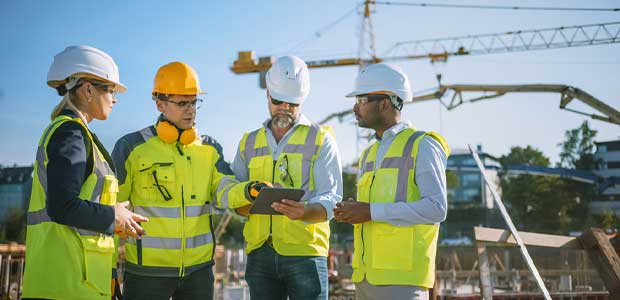 four people in neon vests and hard hats stand together looking at a tablet. There is a crane in the background