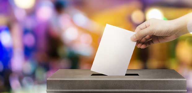 a hand places a piece of paper in a brown ballot box, which is against a blurred purple and yellow background