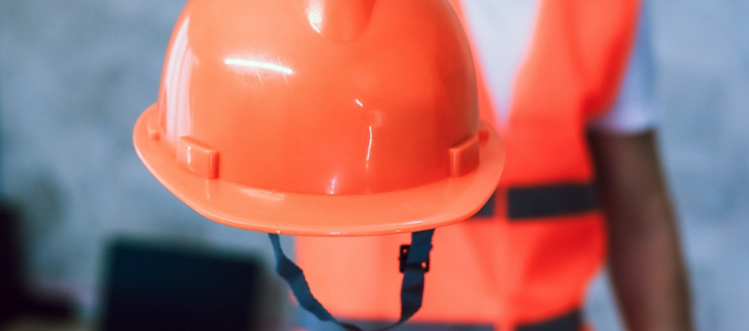 A close-up view of a bright orange construction hard hat featuring a black chin strap, held out by a worker in a high-visibility vest.