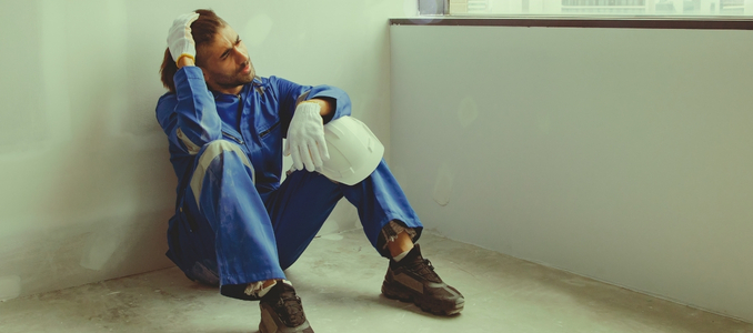 A stressed worker in blue coveralls sitting on the floor against a wall, holding a white hard hat with a hand on his head.