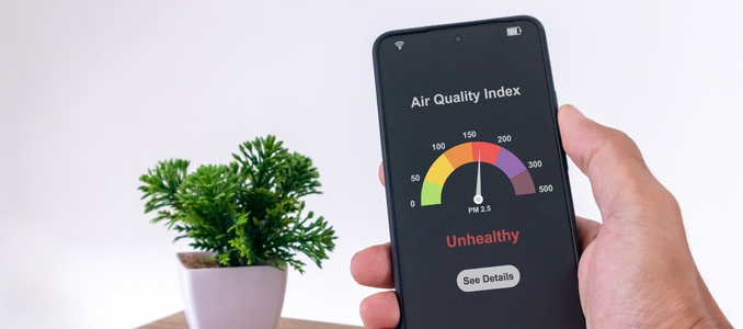 A person holding a smartphone displaying an Air Quality Index app showing a meter pointing to "Unhealthy" with a small potted plant in the background.